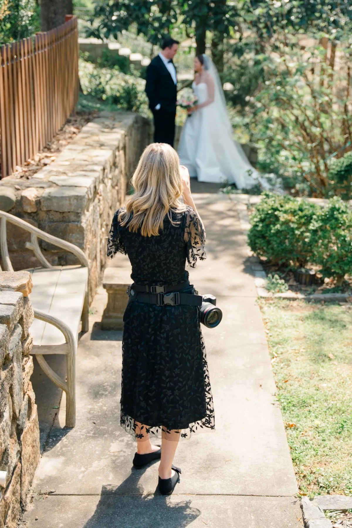 Elizabeth walking down a stone path toward a bride and groom, camera at her hip